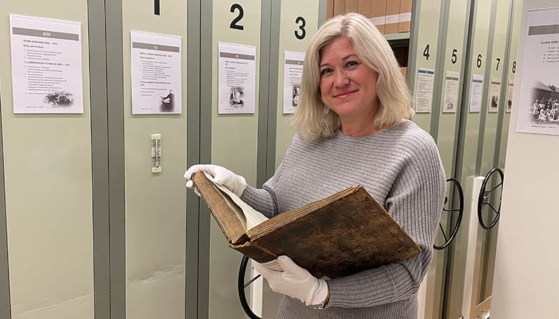 Glad kvinna står i ett arkivrum och håller i en stor bok