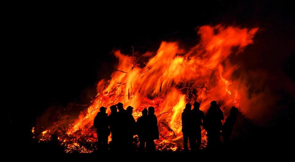 En stor majbrasa brinner. Framför brasan syns siluetter av personer som står och tittar på elden.