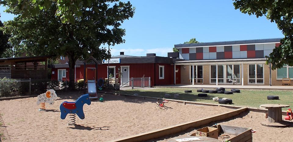 Förskolan Antilopens skolgård. Förskolebyggnaden syns i bakgrunden och på gården är en stor sandlåda med klätterställning och två gungdjur.