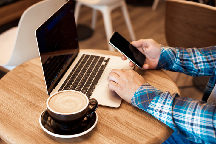 En person sitter med en telefon i handen och har en laptop samt en kopp kaffe framför sig på ett trädbord.