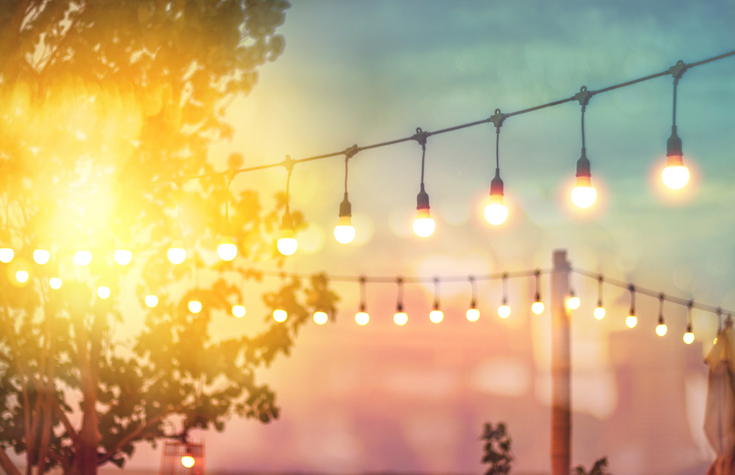 Glödlampor hängandes över en uteplats i solnedgång med ett träd i bakgrunden.