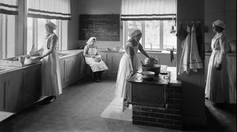 Svartvit bild på fyra flickor med vita förkläden i ett kök som förbereder middagsmat