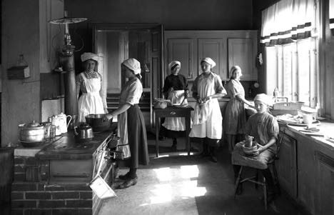 Svartvit bild på sex flickor i förkläden och vita huvuddukar som utför olika matlagningssysslor i köket på skyddshemmet