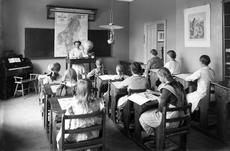 Svartvit bild på 11 flickor som sitter i skolbänkar och lyssnar på lärarinnan som sitter vid katedern längst fram i rummet. 