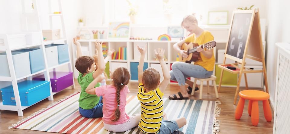 Person sjunger för en grupp med tre barn i ett färgglatt klassrum