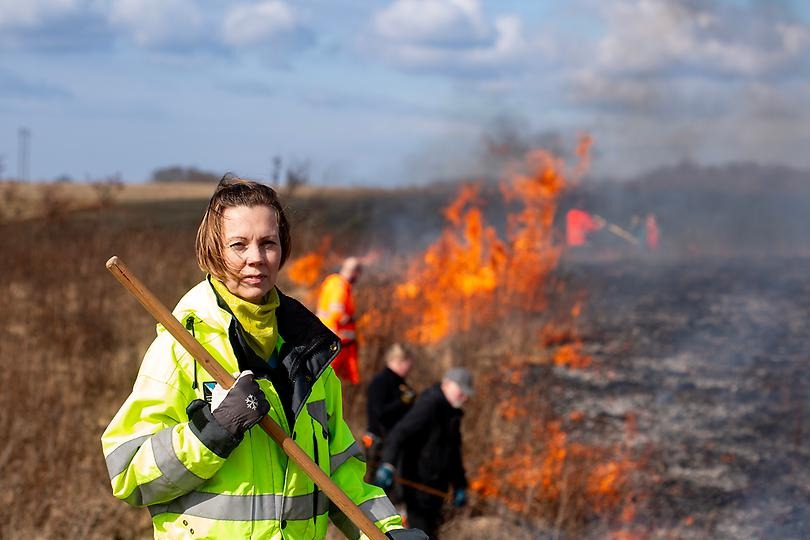 Monica Johansson på ett fält med eldslågor i bakgrunden.