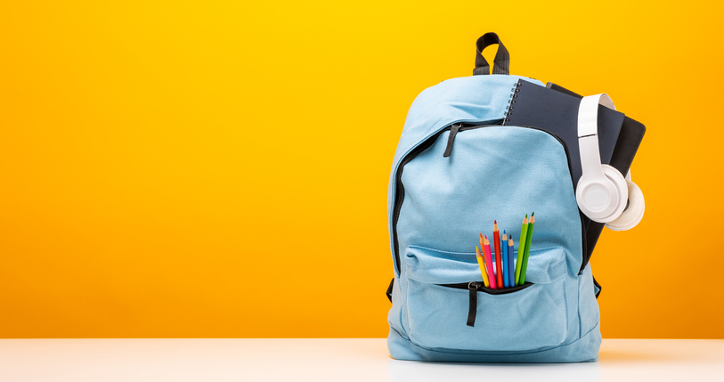 Ljusblå ryggsäck innehållandes pennor som sticker ut ur den samt ett par hörlurar som hänger från den mot orange bakgrund.