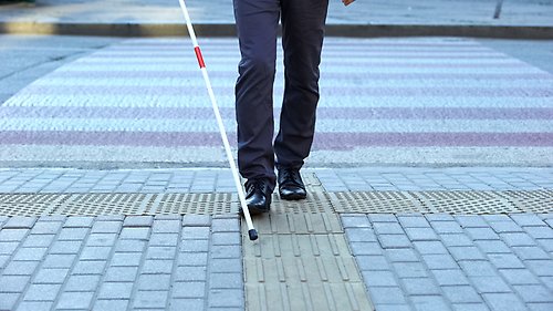 Person med nedsatt syn går över ett övergångsställe samt navigerar sig fram med hjälp av sin blindkäpp.