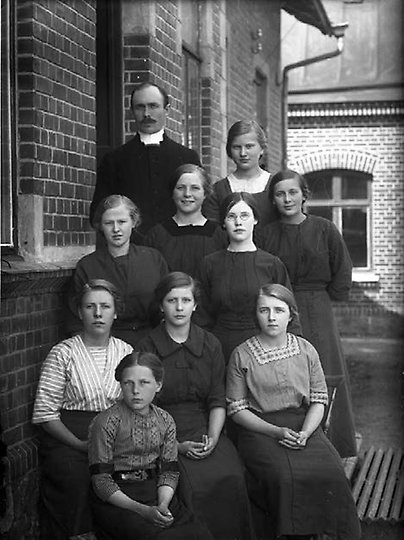 Bild på Komminister Albin Jönsson tillsammans med nio av flickorna som var inskrivna på skyddshemmet 1912. De poserar utanför skyddshemmet. 