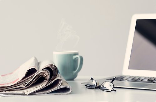 Bild på ett par ihopvikta tidningar på ett bord jämte en turkos kaffekopp samt ett par glasögon framför en laptop. Allt på ett bord.