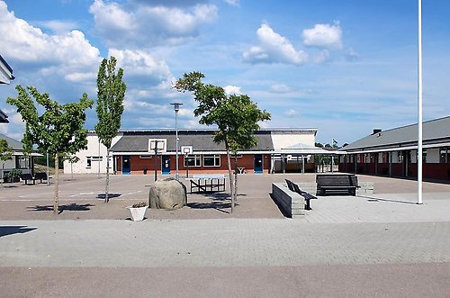 Pilagårdsskolans skolgård. Stor öppen yta för aktiviteter. På ytan finns några träd, bänkar, basketkorgar och en flaggstång.